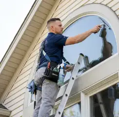 Professional window cleaner on a ladder using a squeegee to clean an exterior arched window, Omaha, NE.